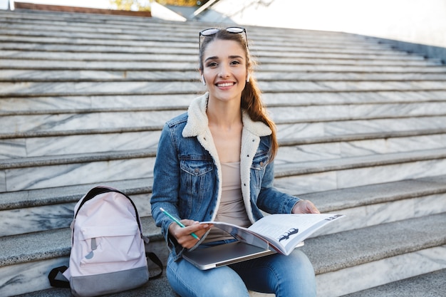 Lachende jonge vrouw draagt jas zittend op de trap buitenshuis, huiswerk, studeren