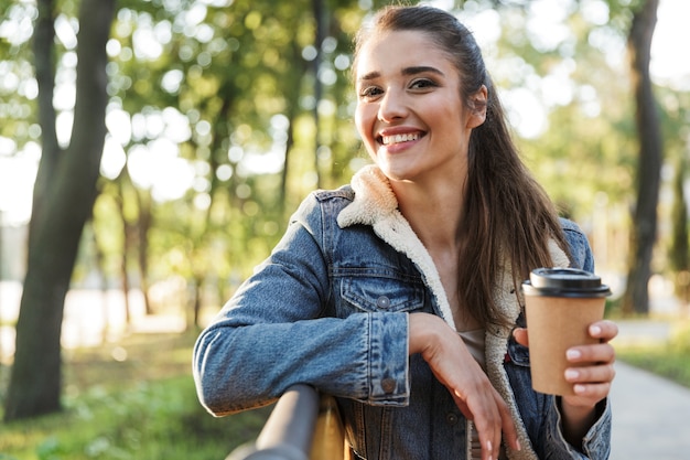 Lachende jonge vrouw draagt jas zittend op een bankje in het park, met afhaalmaaltijden koffiekopje