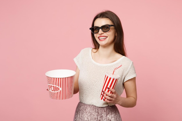 Lachende jonge vrouw in 3d imax bril poseren geïsoleerd op pastel roze achtergrond. Mensen oprechte emoties in de bioscoop, lifestyle concept. Bespotten kopie ruimte. Houd een emmer popcorn, een kopje cola of frisdrank vast.