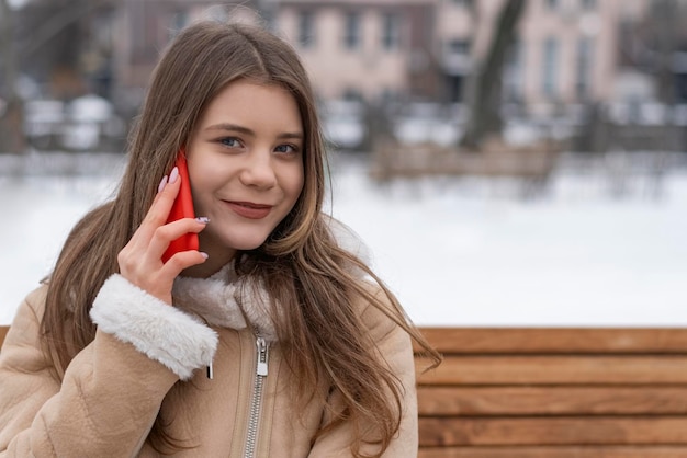 Lachende jonge vrouw in beige schapenvacht jas rust buiten in de winter Schattig meisje zit op een bankje in het park en praat over de telefoon