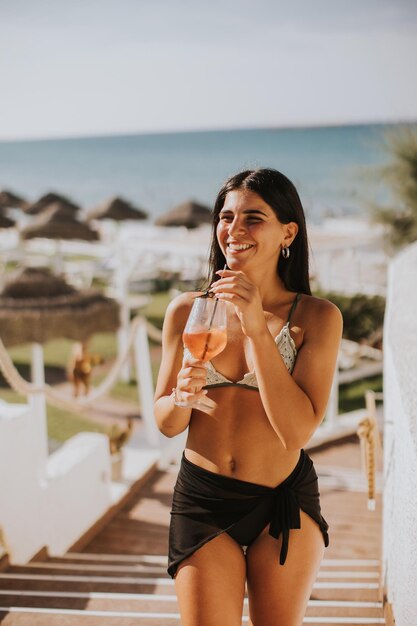 Lachende jonge vrouw in bikini genieten van vakantie op het strand tijdens het drinken van een cocktail