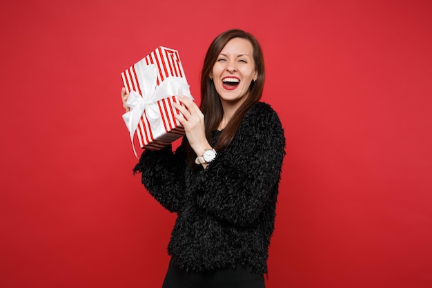 Lachende jonge vrouw in bont trui houdt rood gestreepte huidige doos met cadeau lint geïsoleerd op rode achtergrond. Valentijnsdag, Internationale Vrouwendag, verjaardag, vakantieconcept. Bespotten kopie ruimte.