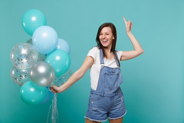 Lachende jonge vrouw in denim kleding wijsvinger omhoog, vieren, kleurrijke luchtballonnen houden geïsoleerd op blauwe turquoise muur achtergrond. Vakantie verjaardagsfeestje, mensen emoties concept.