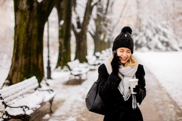 Lachende jonge vrouw in gezellige kleding met behulp van mobiele telefoon en koffiekopje houden op winterdag