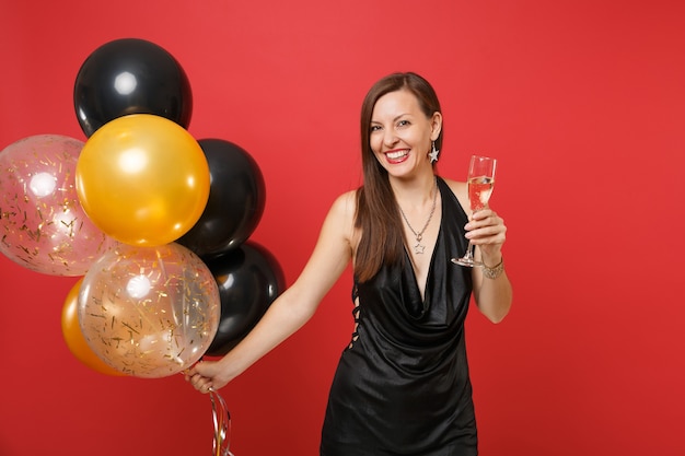 Lachende jonge vrouw in kleine zwarte jurk vieren, met glas champagne, lucht ballonnen geïsoleerd op rode achtergrond. St. Valentijnsdag, Gelukkig Nieuwjaar, verjaardag mockup vakantie feest concept.