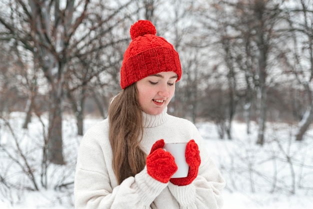 Lachende jonge vrouw in rode gebreide muts en wanten houdt kop hete thee buiten Warme dranken in mok in winterwandeling