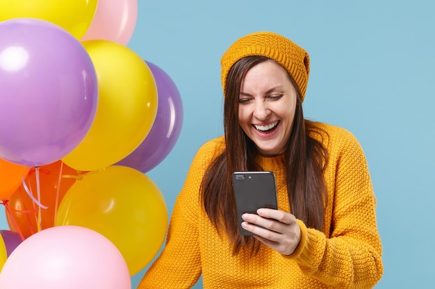 Lachende jonge vrouw in trui hoed poseren geïsoleerd op blauwe achtergrond. Vakantie verjaardagsfeestje, mensen emoties concept. Bespotten kopie ruimte. Vieren houd kleurrijke luchtballonnen vast met behulp van mobiele telefoon.