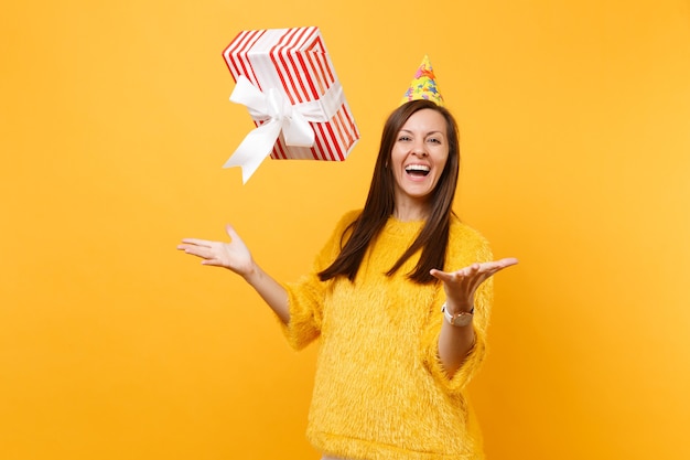 Lachende jonge vrouw in verjaardagsfeestje hoed overgeven rode doos met cadeau aanwezig, vieren en genieten van vakantie geïsoleerd op heldere gele achtergrond. mensen oprechte emoties, lifestyle concept.