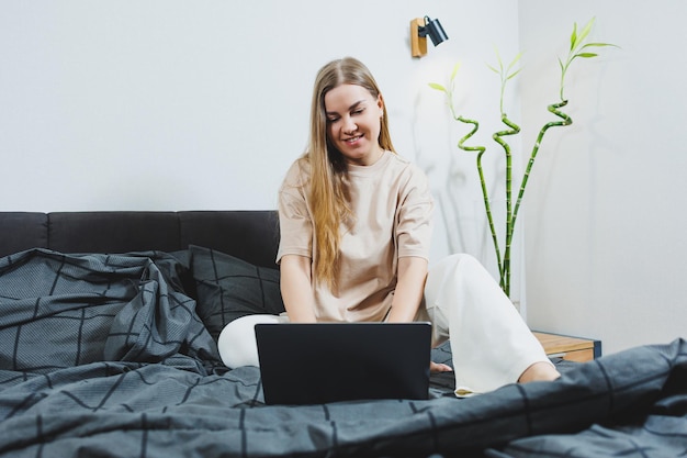 Lachende jonge vrouw in vrijetijdskleding glimlachend en op afstand werkend zittend op bed met laptop tijdens werk op afstand Freelance werk Werk vanuit huis
