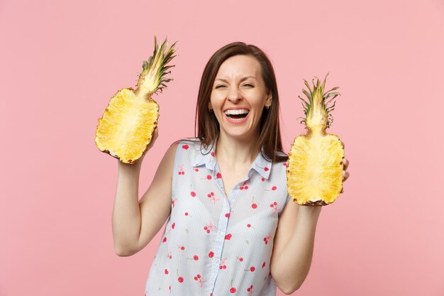 Lachende jonge vrouw in zomerkleren met helften van vers rijp ananasfruit geïsoleerd op roze pastelmuurachtergrond in studio. Mensen levendige levensstijl ontspannen vakantie concept. Bespotten kopie ruimte.