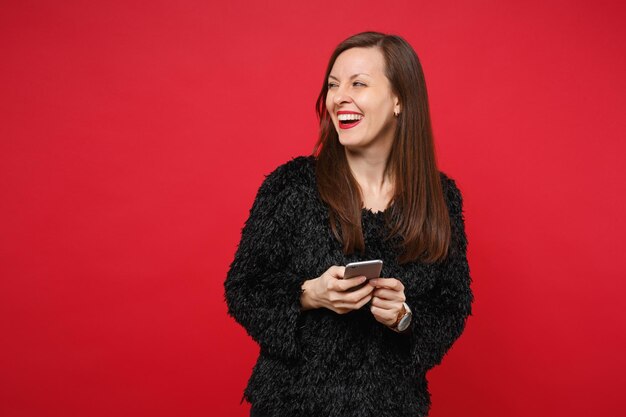 Lachende jonge vrouw in zwarte vacht trui opzij kijken, met behulp van mobiele telefoon geïsoleerd op heldere rode muur achtergrond in studio. mensen oprechte emoties, lifestyle concept. bespotten kopie ruimte.