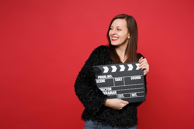 Lachende jonge vrouw in zwarte vacht trui opzij kijken, met klassieke zwarte film filmklapper geïsoleerd op rode muur achtergrond. mensen oprechte emoties, lifestyle concept. bespotten kopie ruimte.