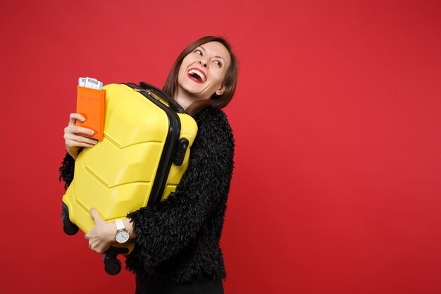 Lachende jonge vrouw in zwarte vacht trui opzoeken, met koffer, paspoort instapkaart ticket geïsoleerd op heldere rode achtergrond. Mensen oprechte emoties, lifestyle concept. Bespotten kopie ruimte.