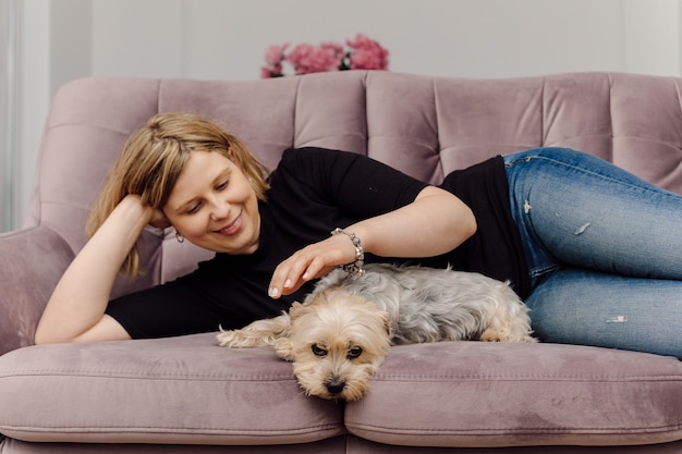 Lachende jonge vrouw ligt op roze sofa in de woonkamer, Yorkshire Terrier aaien.