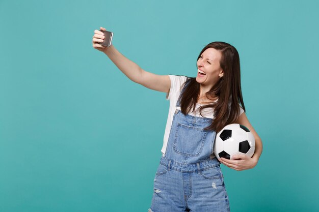 Lachende jonge vrouw voetbalfan met voetbal doen selfie schot op mobiele telefoon geïsoleerd op blauwe turkooizen achtergrond. Mensen emoties, sport familie leisure lifestyle concept. Bespotten kopie ruimte.