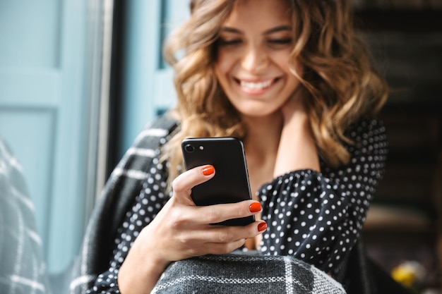 Lachende jonge vrouw zitten gewikkeld in een deken bij het raam, met behulp van mobiele telefoon