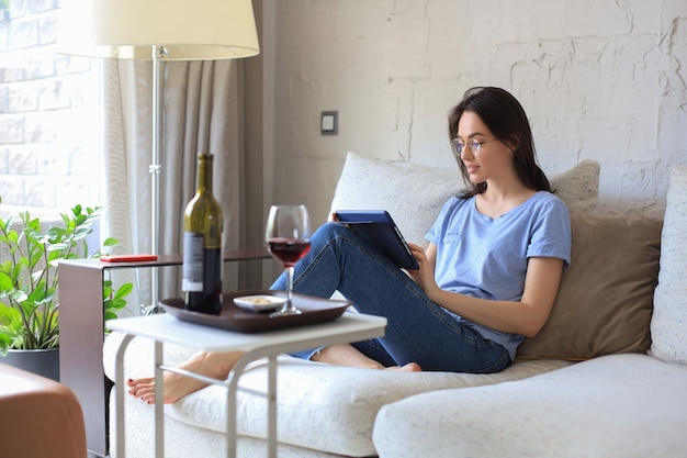 Lachende jonge vrouw zittend op de bank met digitale tablet en chatten met vrienden, wijn drinken.