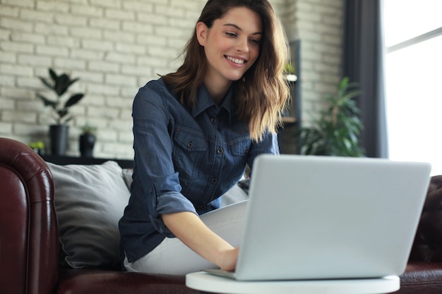 Lachende jonge vrouw zittend op de bank met laptopcomputer en chatten met vrienden.