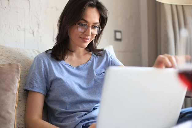 Lachende jonge vrouw zittend op de bank met laptopcomputer en chatten met vrienden.