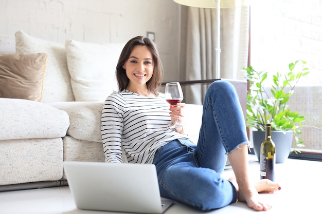 Lachende jonge vrouw zittend op de vloer met laptopcomputer en chatten met vrienden, wijn drinken.