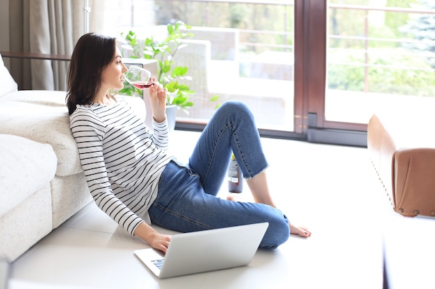 Lachende jonge vrouw zittend op de vloer met laptopcomputer en chatten met vrienden, wijn drinken.