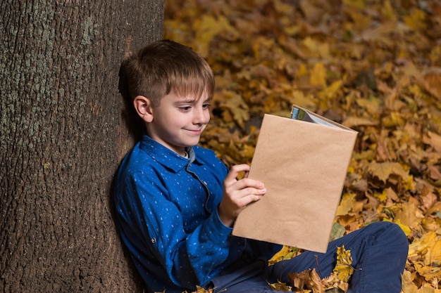 Lachende jongen zit in de buurt van boom in herfst bos en leesboek. ruimte kopiëren. spotten.