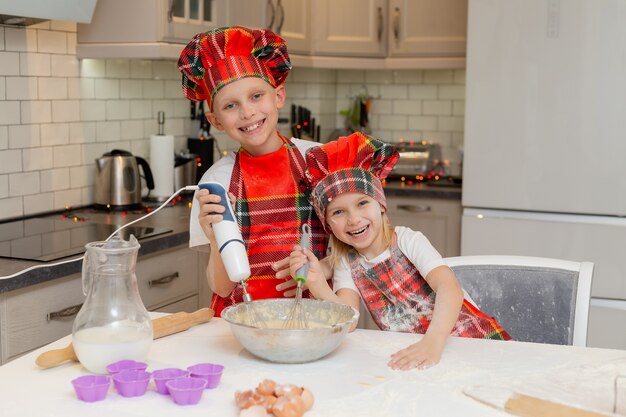 lachende kinderen in chef-kokkostuums bereiden deeg van meelmelkeieren voor kerstgebakjes