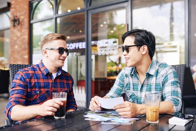 Lachende knappe jonge mannen in zonnebril zitten aan café tafel en gedrukte foto's van zomervakantie bespreken