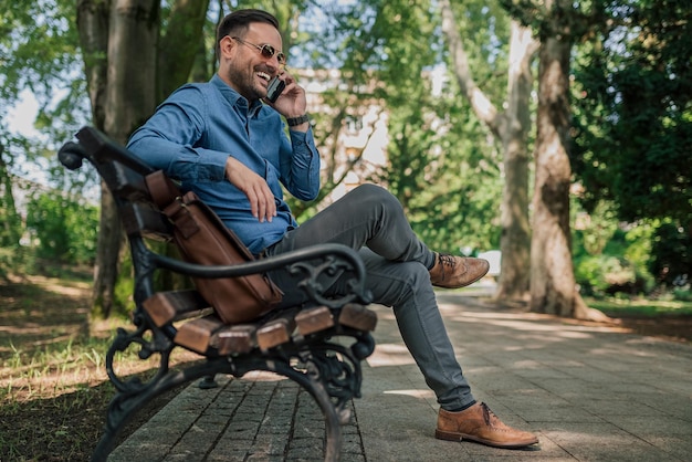 Lachende knappe ondernemer praten op slimme telefoon zittend op bankje in het park