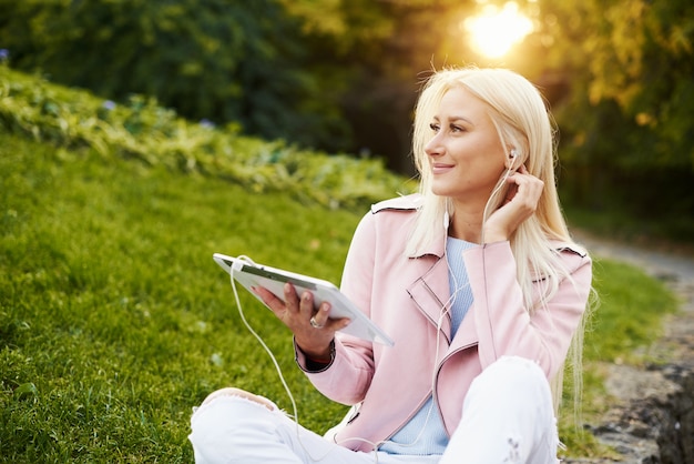 Lachende knappe vrouw luisteren ontspannende muziek in de koptelefoon met haar mobiele telefoon of tablet in het park. student luistert naar een online college en bereidt zich voor op tentamens.