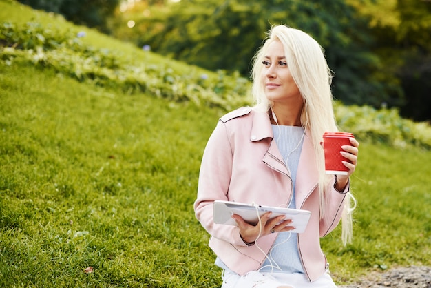 Lachende knappe vrouw luisteren ontspannende muziek in de koptelefoon met haar mobiele telefoon of tablet in het park. student luistert naar een online college en bereidt zich voor op tentamens.