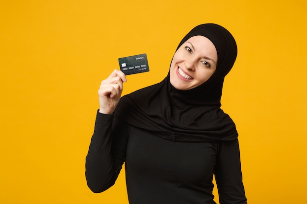 Lachende leuke jonge arabische moslimvrouw in hijab zwarte kleding houd in de hand creditcard geïsoleerd op gele muur portret. mensen religieuze levensstijl concept.