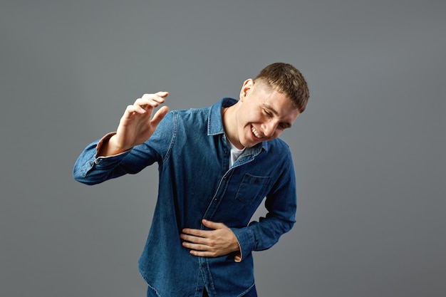 Lachende man gekleed in een spijkerbroek houdt zijn hand op de buik in de studio op de grijze achtergrond.