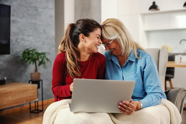 Lachende moeder en dochter die samen thuis zitten en laptop gebruiken om op sociale media te hangen