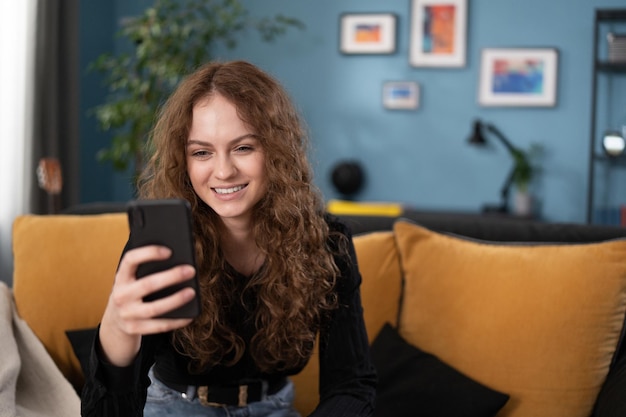 Lachende mooie brunette tienermeisje met krullend haar gebruikt smartphone terwijl ze op de bank zit