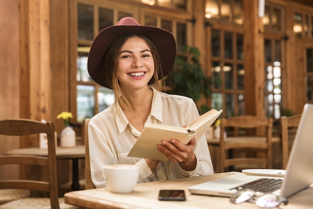 Lachende mooie vrouw in hoed zit aan de tafel van het café binnenshuis