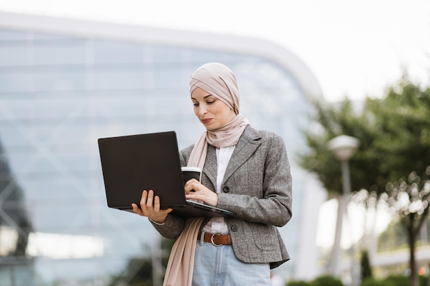 Lachende moslimdame in hijab die zich voordeed op de camera terwijl ze aan een laptop buiten kantoor werkte