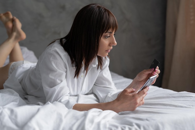 Lachende, ontspannen jonge vrouw die op het bed ligt, leest haar creditcardnummer en voert op een smartphone aankopen op internet uit en betaalt gemakkelijk veilig thuisbankieren