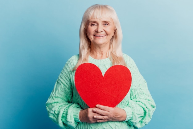 Lachende oudere vrouw houdt rood hart kaart geïsoleerd op blauwe achtergrond