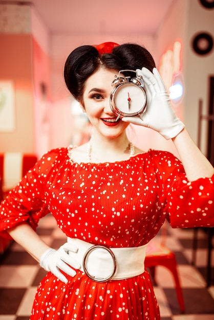 Foto lachende pin-up girl poseren met wekker, vintage café-interieur, populaire amerikaanse mode 50s en 60s. rode jurk met stippen, lichte make-up