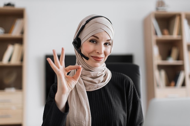 Lachende positieve zelfverzekerde vrouw in hijab en headset zittend aan een bureau