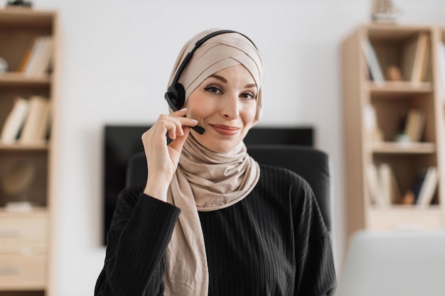 Lachende positieve zelfverzekerde vrouw in hijab en headset zittend aan een bureau