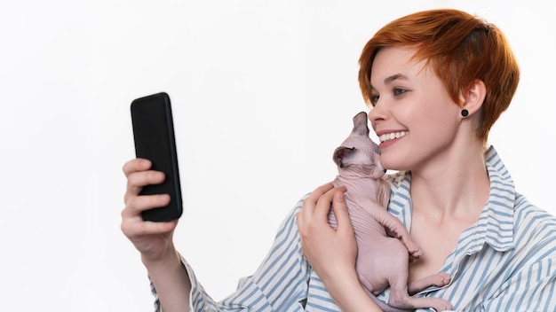 Lachende roodharige jonge vrouw knuffelen Sphynx Cat neemt selfie foto op smartphone