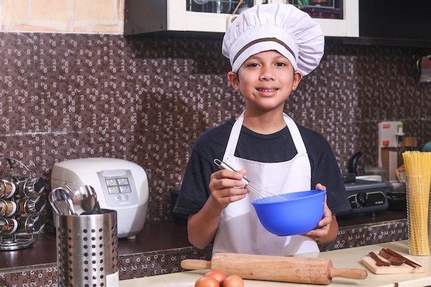 Lachende schattige jongen in chef-kok uniform deeg maken op kom met behulp van garde tegen keukenachtergrond