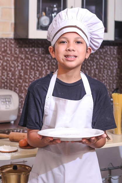 Lachende schattige jongen in chef-kok uniform met lege witte plaat of schotel in de keuken