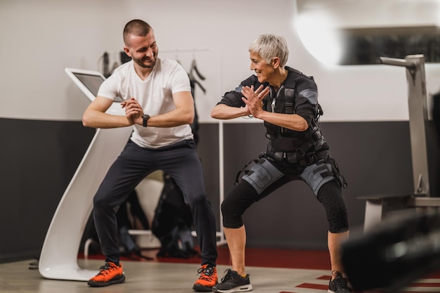 Lachende senior vrouw doet EMS trainen met personal trainer in de sportschool.