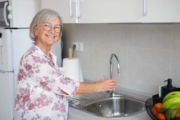 Lachende senior vrouw kraanwater drinken uit glas in de keuken