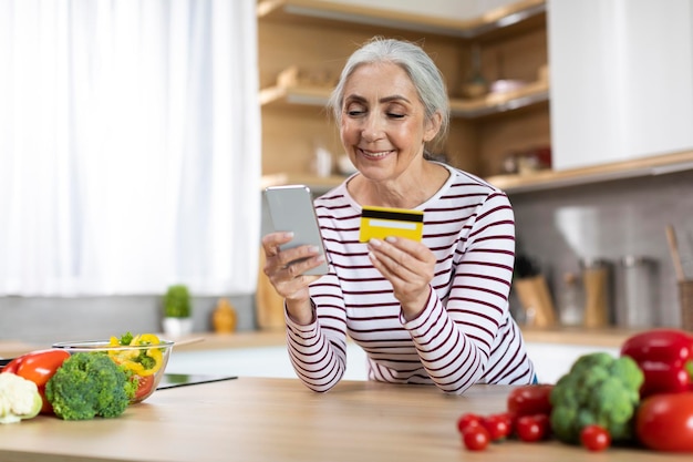 Lachende Senior vrouw met behulp van smartphone en creditcard tijdens het koken in de keuken