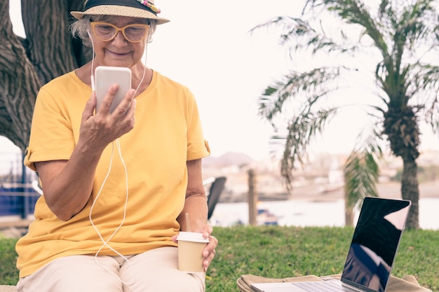 Lachende senior vrouw met koptelefoon buiten zitten met behulp van slimme telefoon met een koffiekopje. Laptopcomputer dicht bij haar