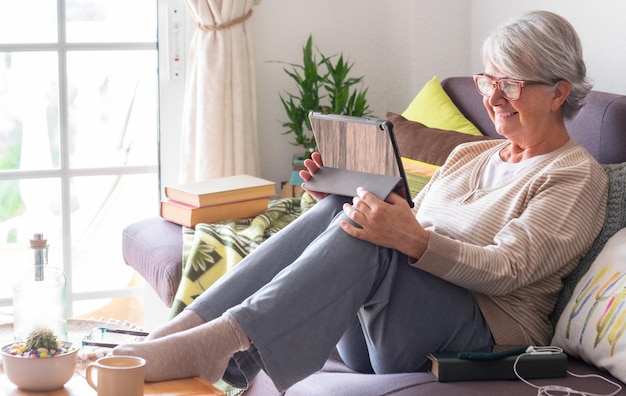 lachende senior vrouw met wit haar ontspant thuis liggend op de bank kijkend naar haar digitale tablet